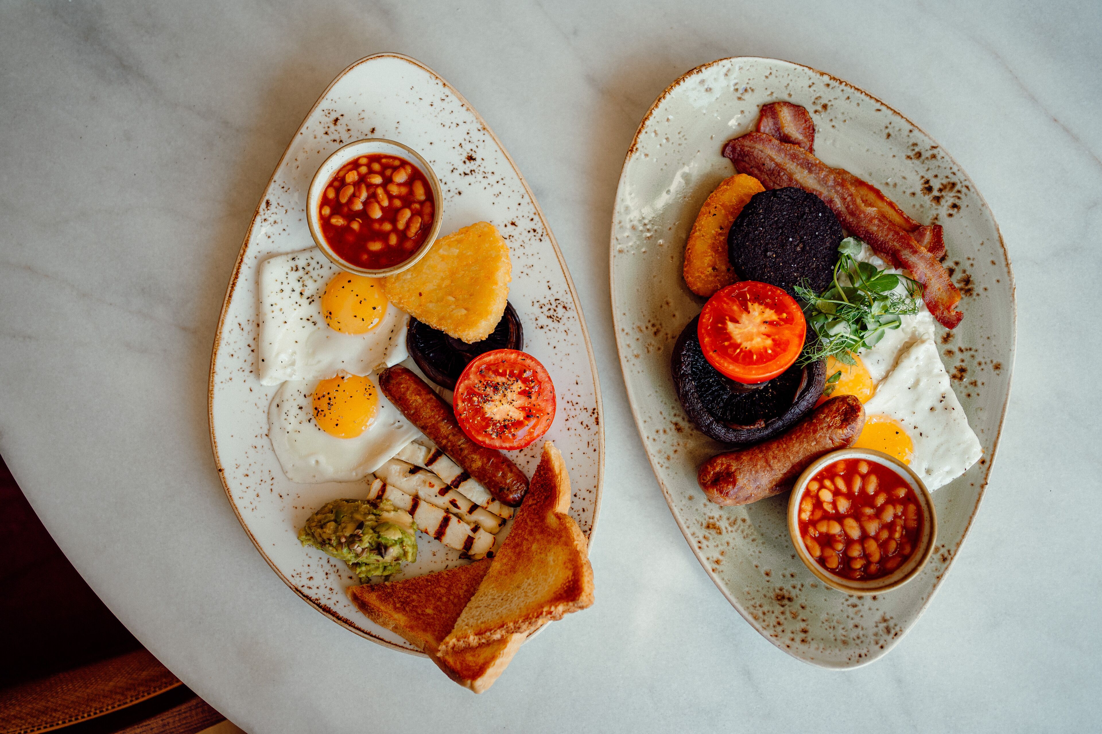 Two plates of breakfast with eggs, sausages, bacon, toast, and beans.