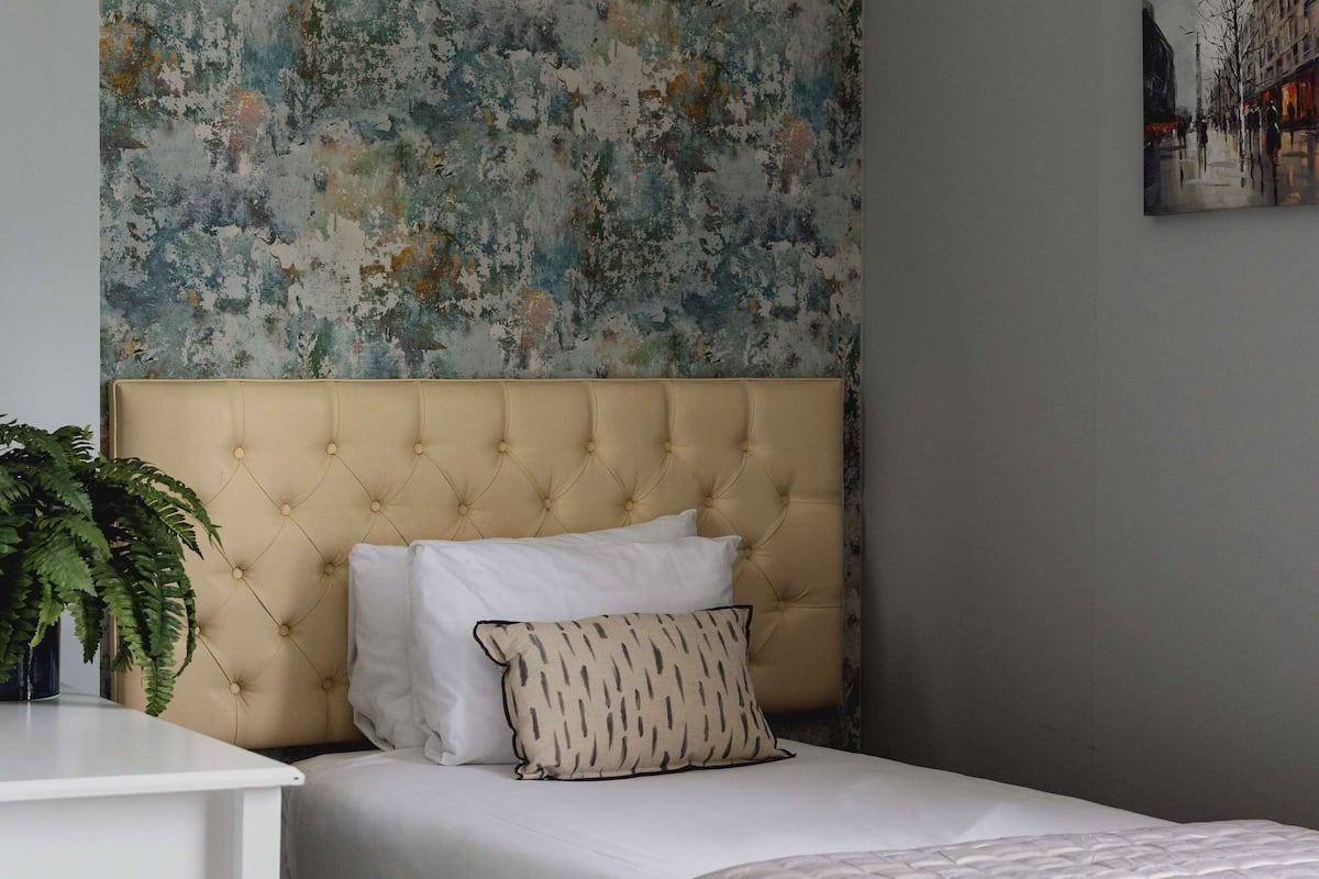 A bedroom with a tufted headboard, a bed with white bedding, a patterned pillow, a small bedside table with a plant, and a wall with a floral wallpaper.