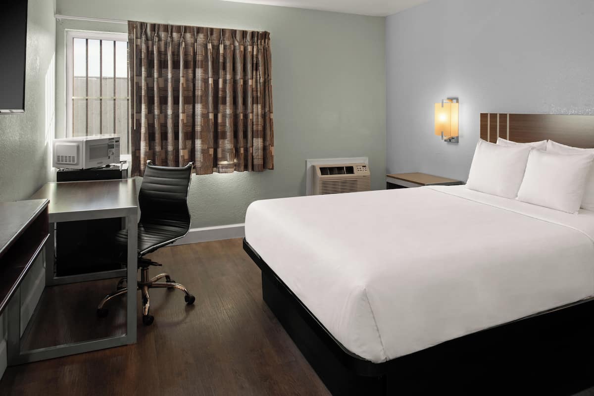 A hotel room with a bed, desk, chair, microwave, air conditioner, and a window with curtains.