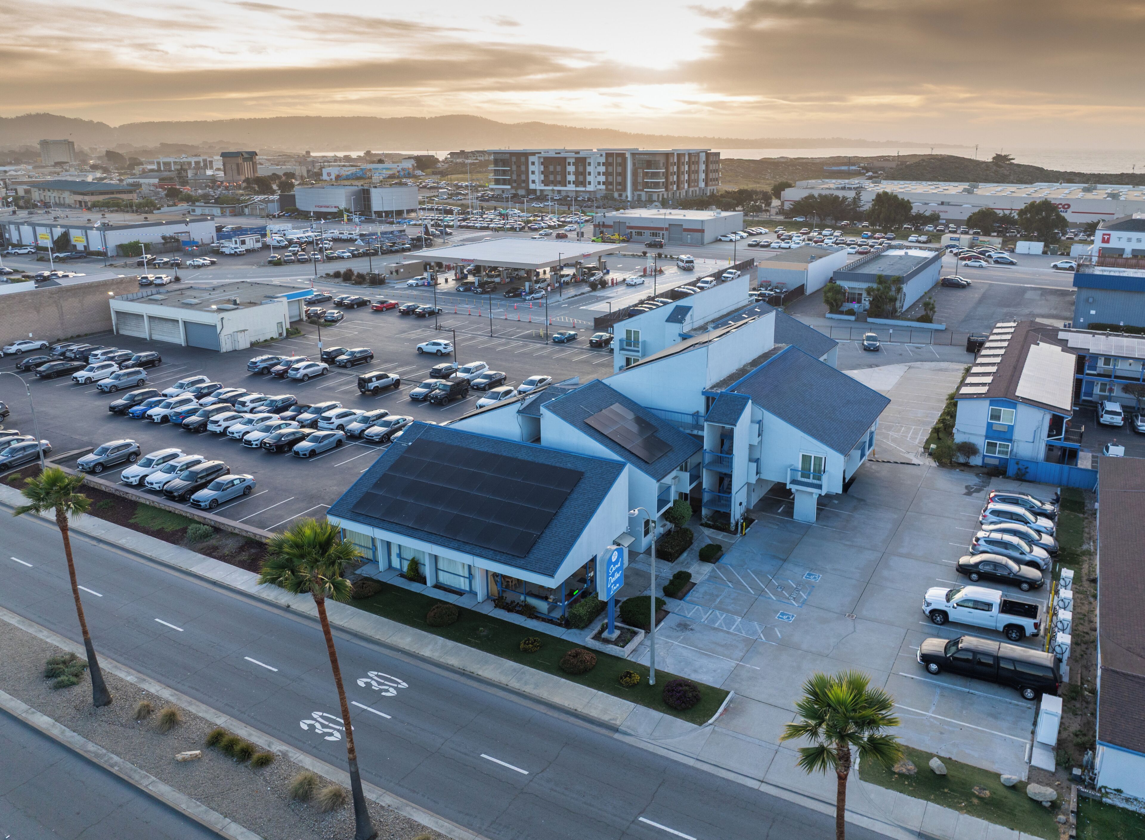 A parking lot with multiple cars, a building with a blue roof, and a view of a hilly landscape in the background.