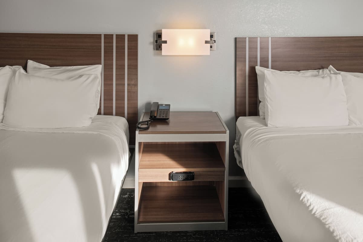 A hotel room with two beds, a nightstand with a phone and a drawer, and a wall-mounted light fixture.
