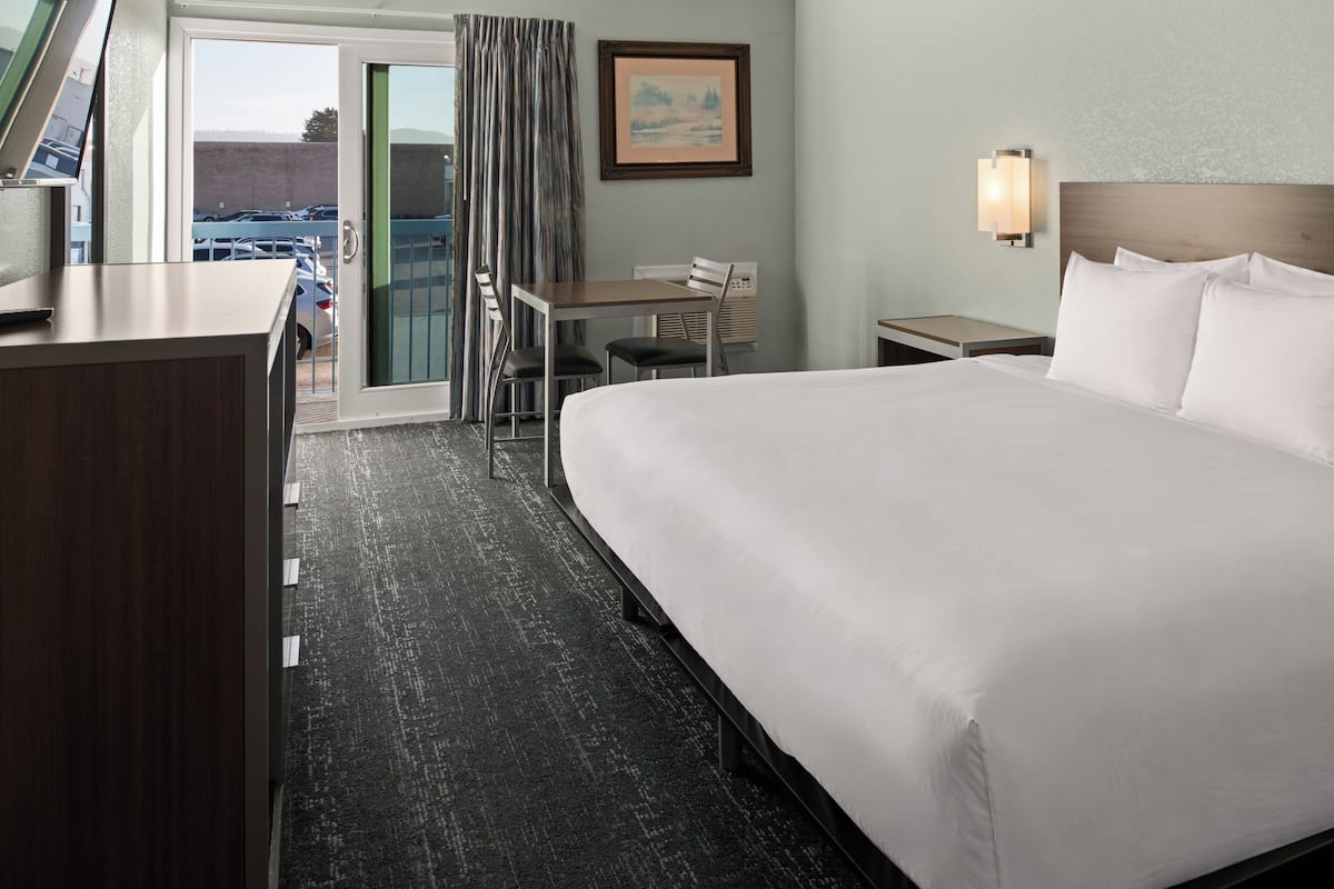 A hotel room with a large bed, a desk, a chair, a small table, a nightstand, a wall-mounted lamp, and a view of parked cars through a glass door.
