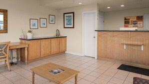 A kitchen with wooden cabinets, a sink, and a countertop. There are framed pictures on the wall, a door, and a bulletin board with notices. - Sand Dollar Inn (Seaside)