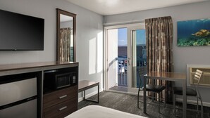 A hotel room with a bed, a desk, a chair, a TV, a microwave, a dresser, a balcony with a view, and a painting of a turtle. - Sand Dollar Inn (Seaside)
