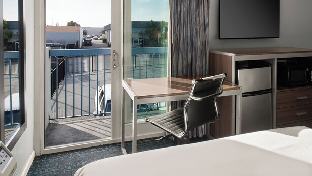 A hotel room with a bed, desk, chair, TV, and a balcony with a view of the parking lot and buildings. - Sand Dollar Inn (Seaside)