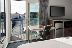 A hotel room with a bed, desk, chair, TV, and a balcony with a view of the parking lot and buildings.