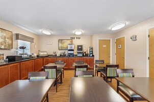 A hotel dining area with wooden tables and chairs, a kitchen area with appliances, and a clock on the wall.