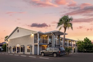 Un motel de dos pisos con un balcón en el nivel superior, un coche estacionado al frente y una palmera a la derecha.