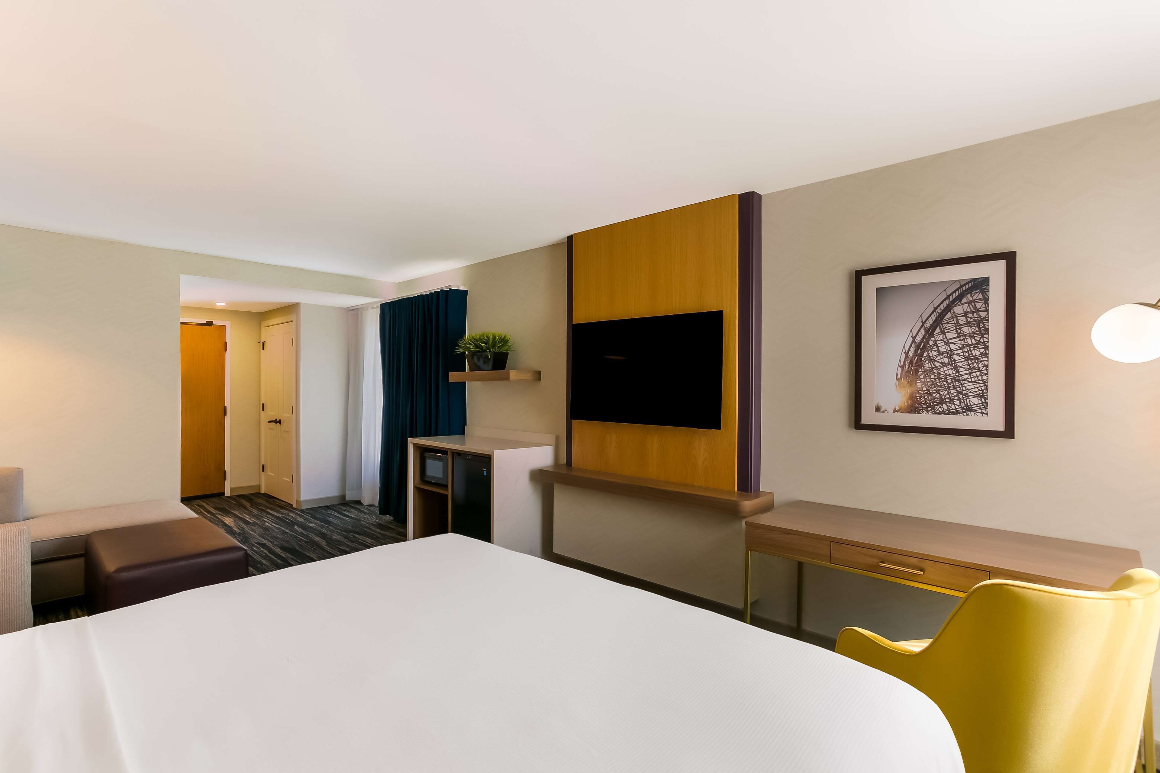A hotel room with a bed, desk, chair, TV, and artwork on the wall.
