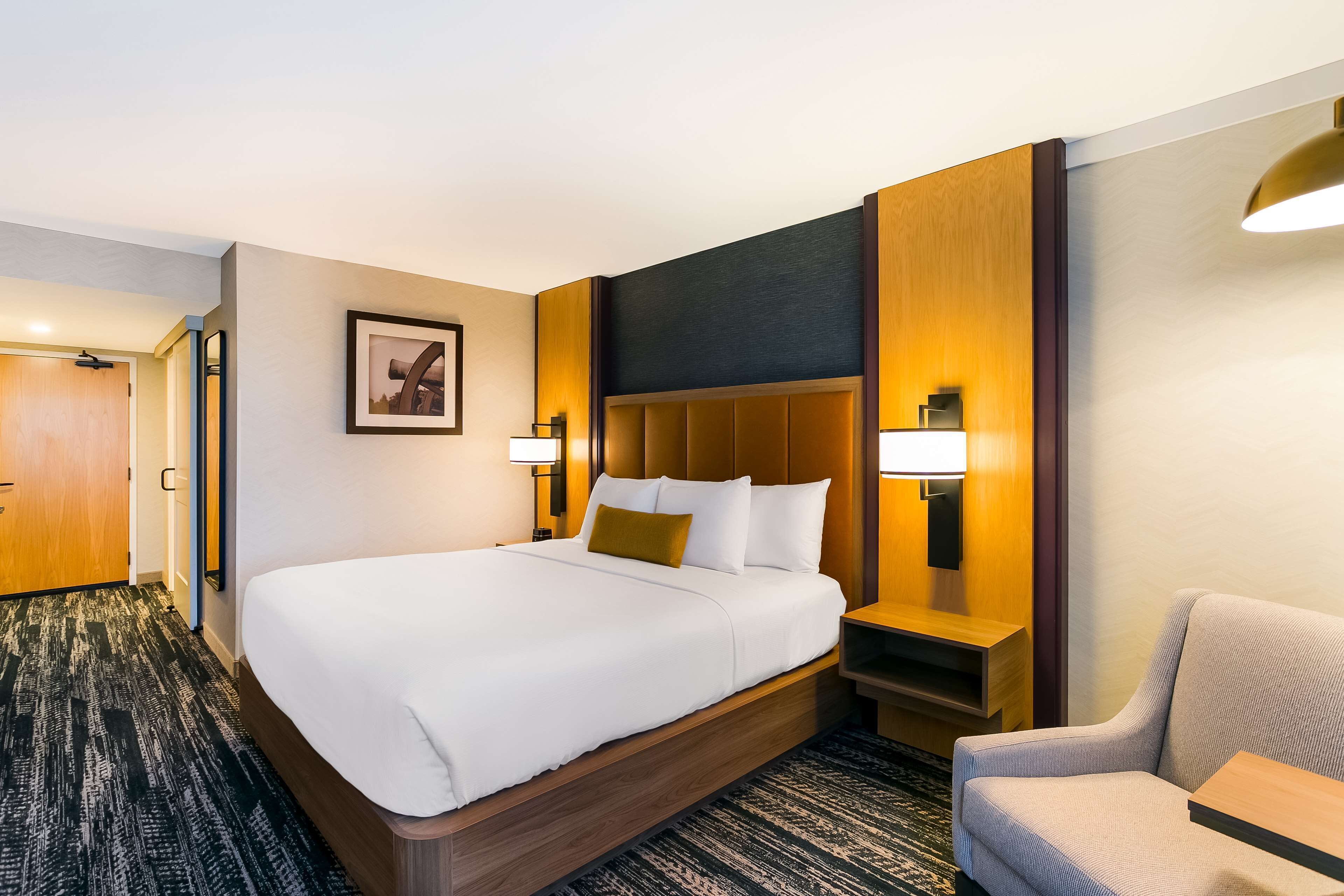 A hotel room with a large bed, a wooden headboard, a nightstand, a lamp, a chair, and a carpeted floor.