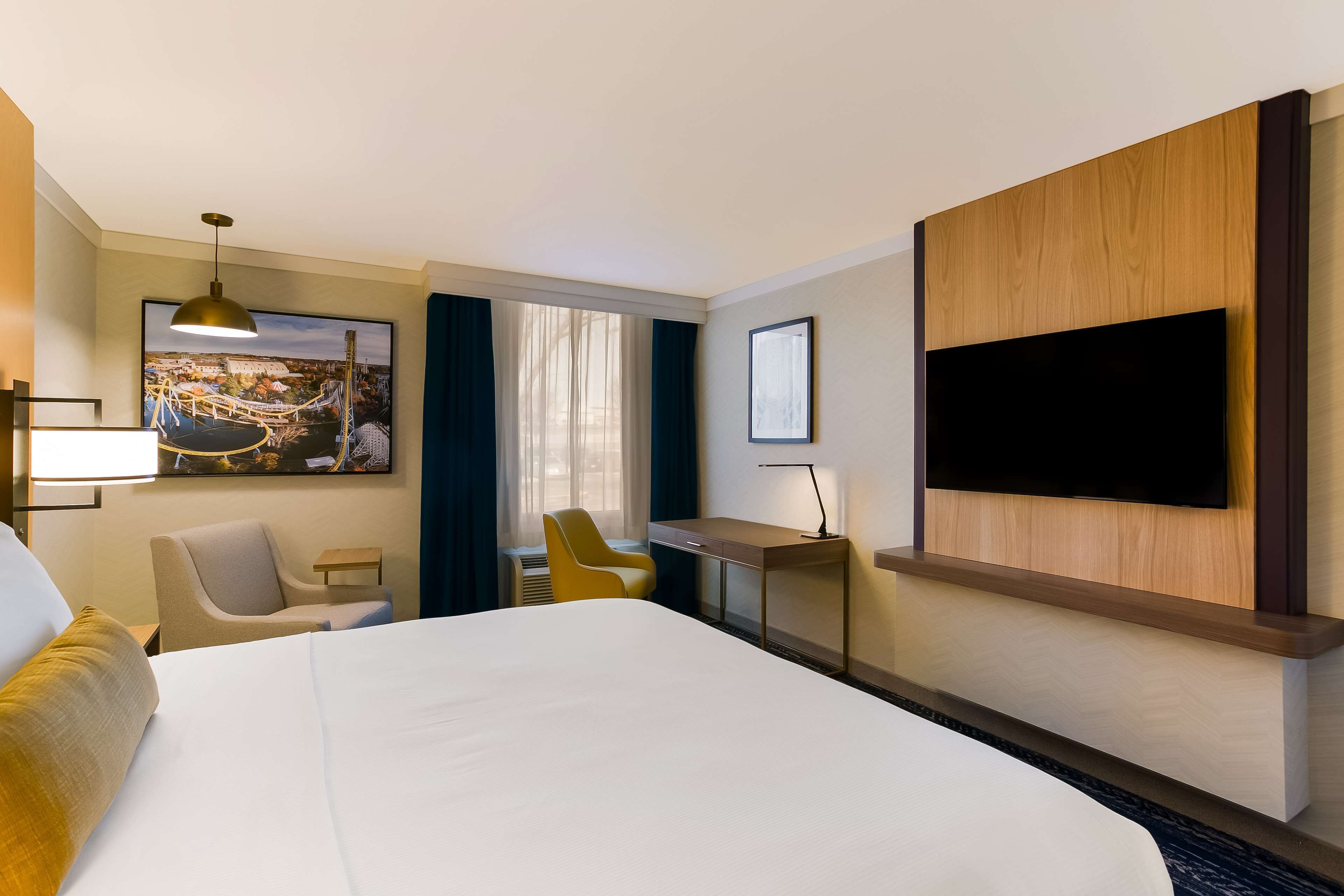 A hotel room with a large bed, a desk, a chair, a television mounted on the wall, and a window with curtains.