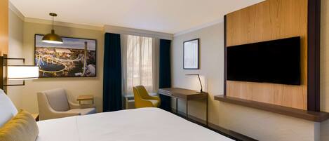 A hotel room with a large bed, a desk, a chair, a television mounted on the wall, and a window with curtains.