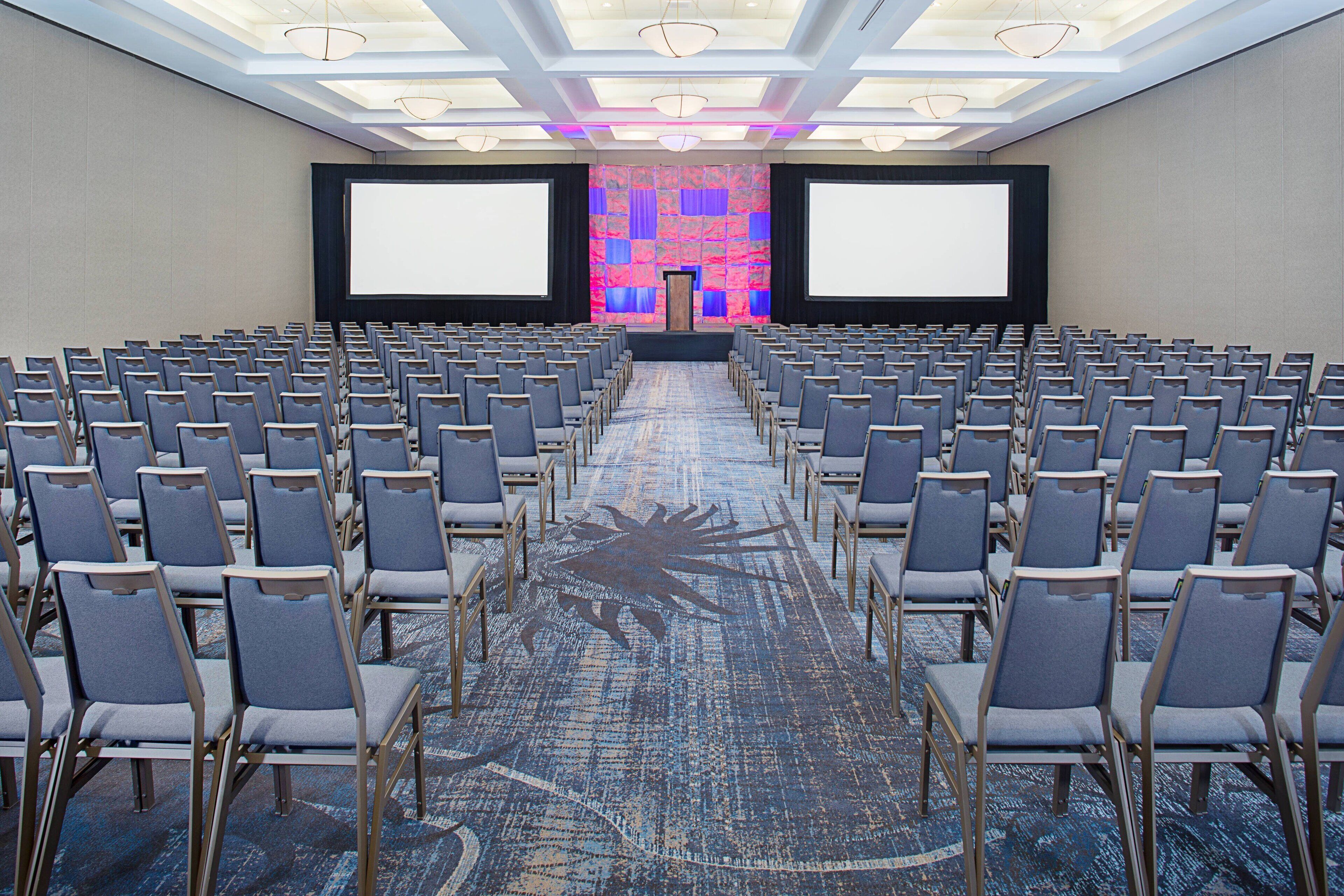 A conference room with rows of chairs, a stage with a large screen, and a patterned carpet.