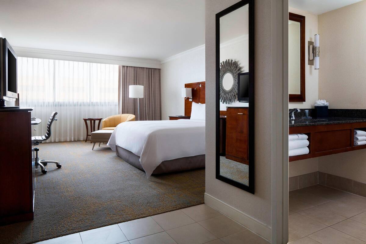Habitación de hotel con cama, escritorio con silla, televisor, espejo grande y baño con lavamanos y toallero.
