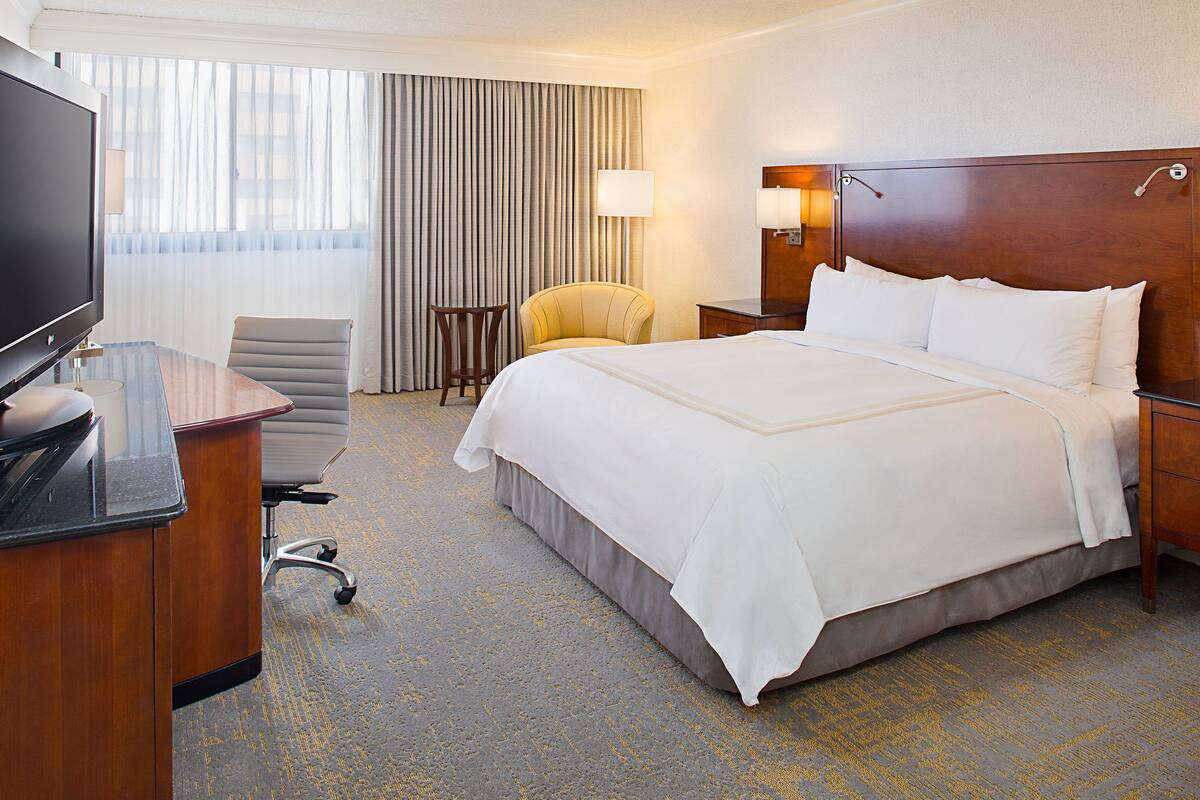 Habitación de hotel con una cama grande, un escritorio con televisor de pantalla plana, una silla, una lámpara y una ventana con cortinas.