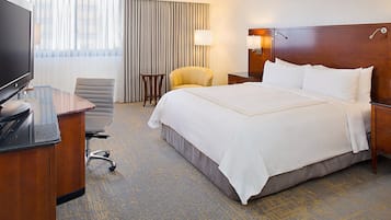 A hotel room with a large bed, a desk with a flat-screen TV, a chair, a lamp, and a window with curtains.