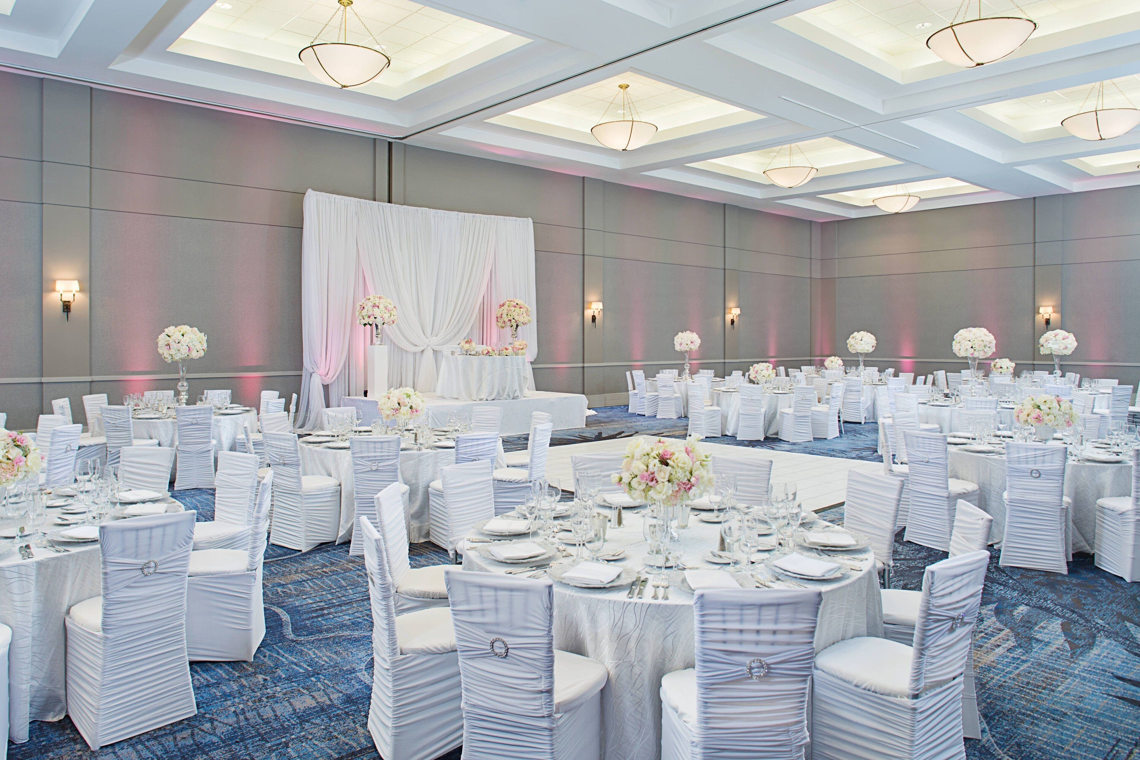 A large banquet hall with round tables set for events, white drapery, and floral centerpieces.