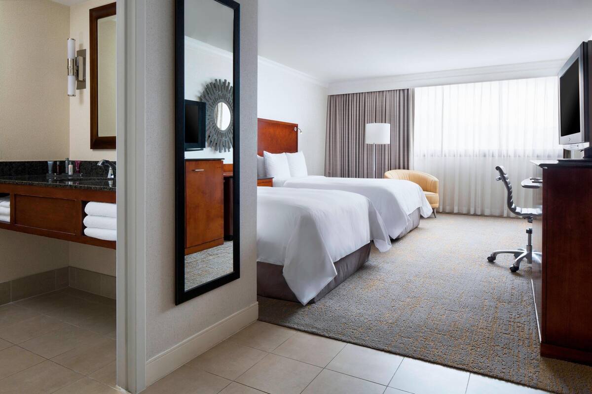 Habitación de hotel con dos camas, baño con lavamanos y espejo, televisor y escritorio.