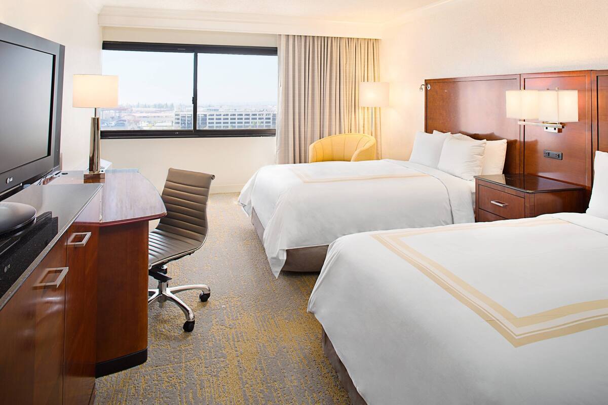 Habitación de hotel con dos camas, un escritorio con televisor, una silla y vista a la ciudad desde la ventana.