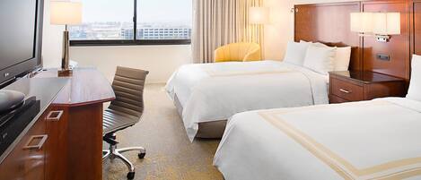 A hotel room with two beds, a desk with a TV, a chair, and a view of the city through the window.