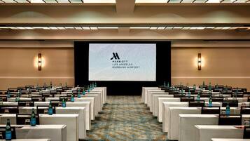 A conference room with rows of tables, each with a name card holder and water bottle. A large screen at the front displays the Marriott logo.