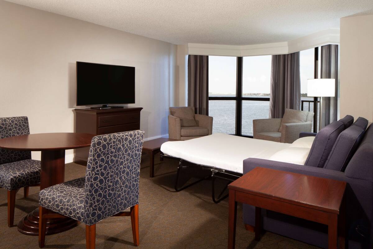 A hotel room with a bed, two armchairs, a sofa, a TV, and a view of the water.
