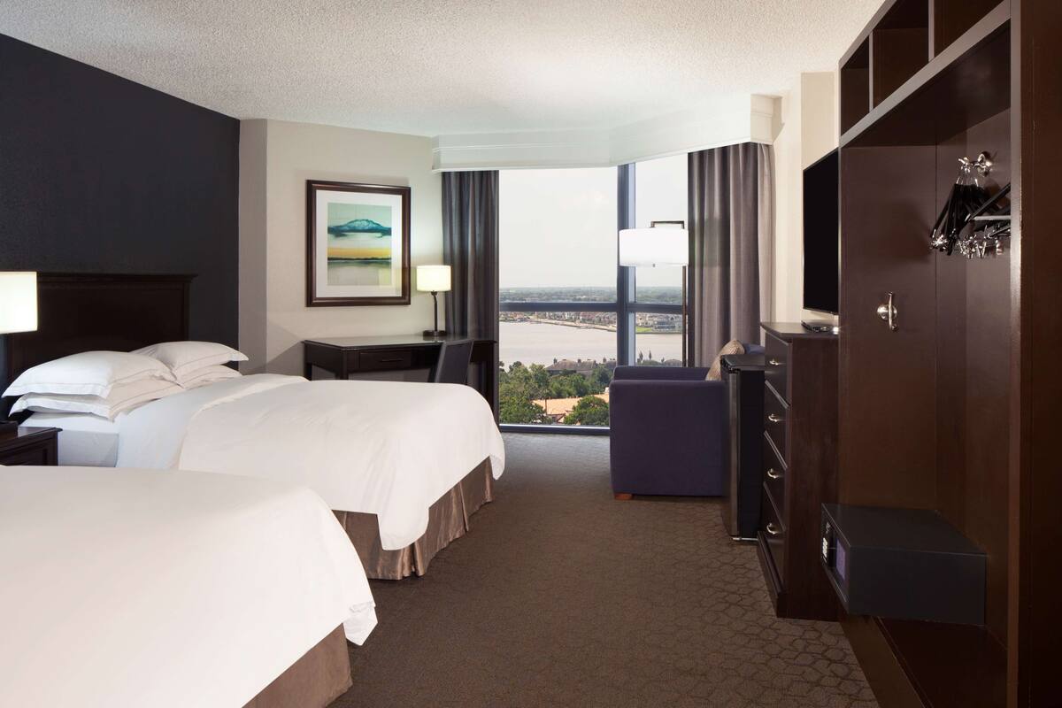 A hotel room with two beds, a desk, a chair, a wardrobe, and a large window offering a city view.