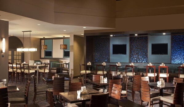 A restaurant with multiple dining tables and chairs, recessed lighting, and a patterned wall with two flat-screen TVs. - Hilton Houston NASA Clear Lake (Houston)