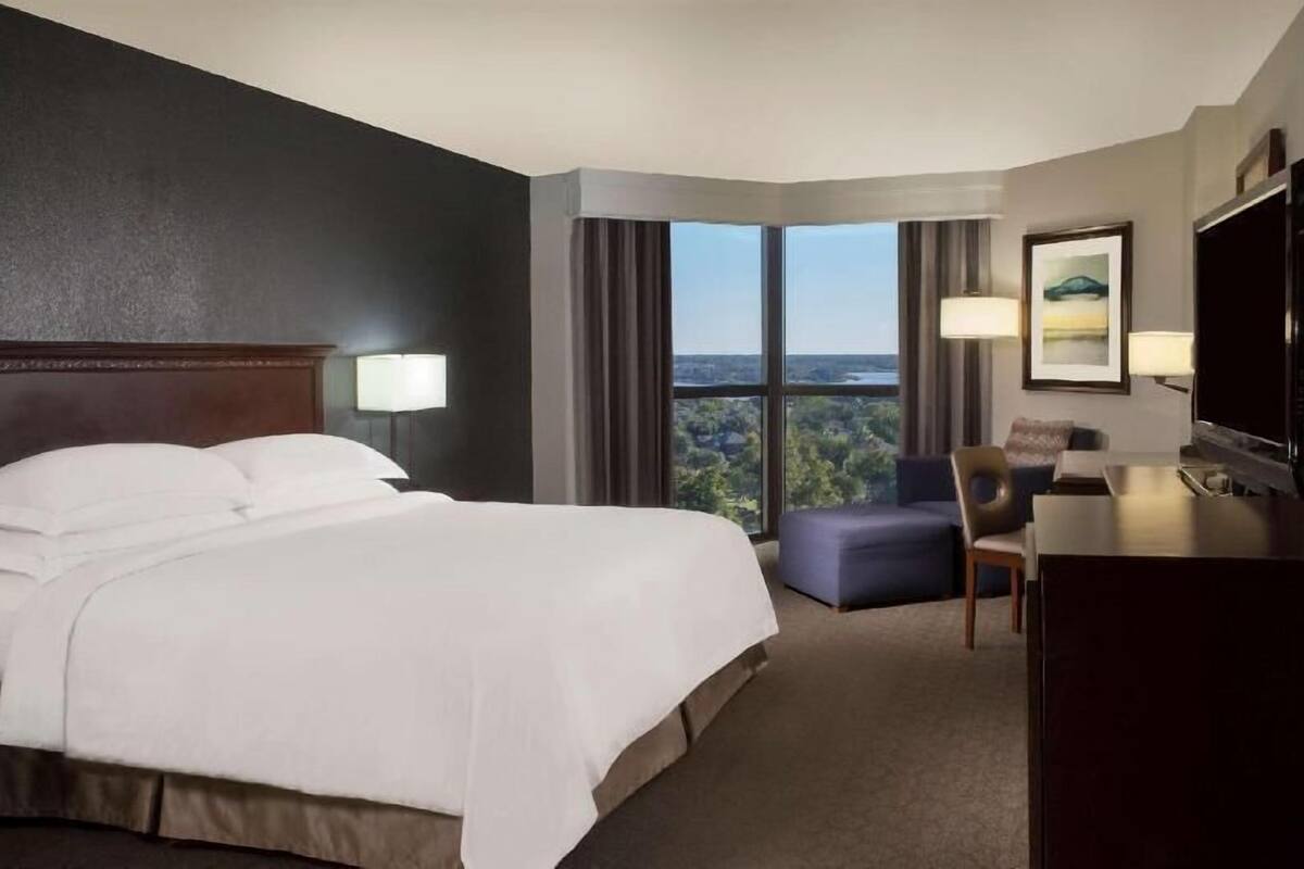 A hotel room with a large bed, a desk, a chair, a TV, and a window with a view.