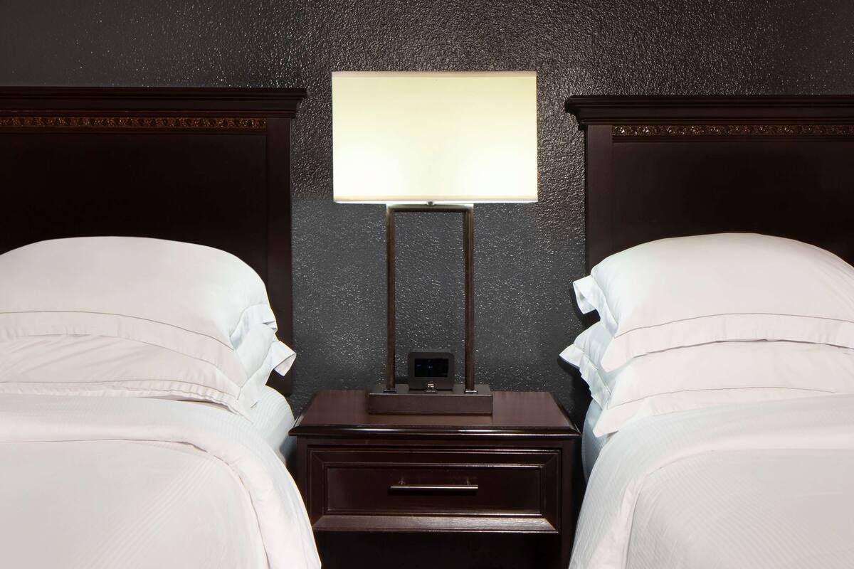 A hotel room with two beds, a nightstand with a lamp, and a dark textured wall.