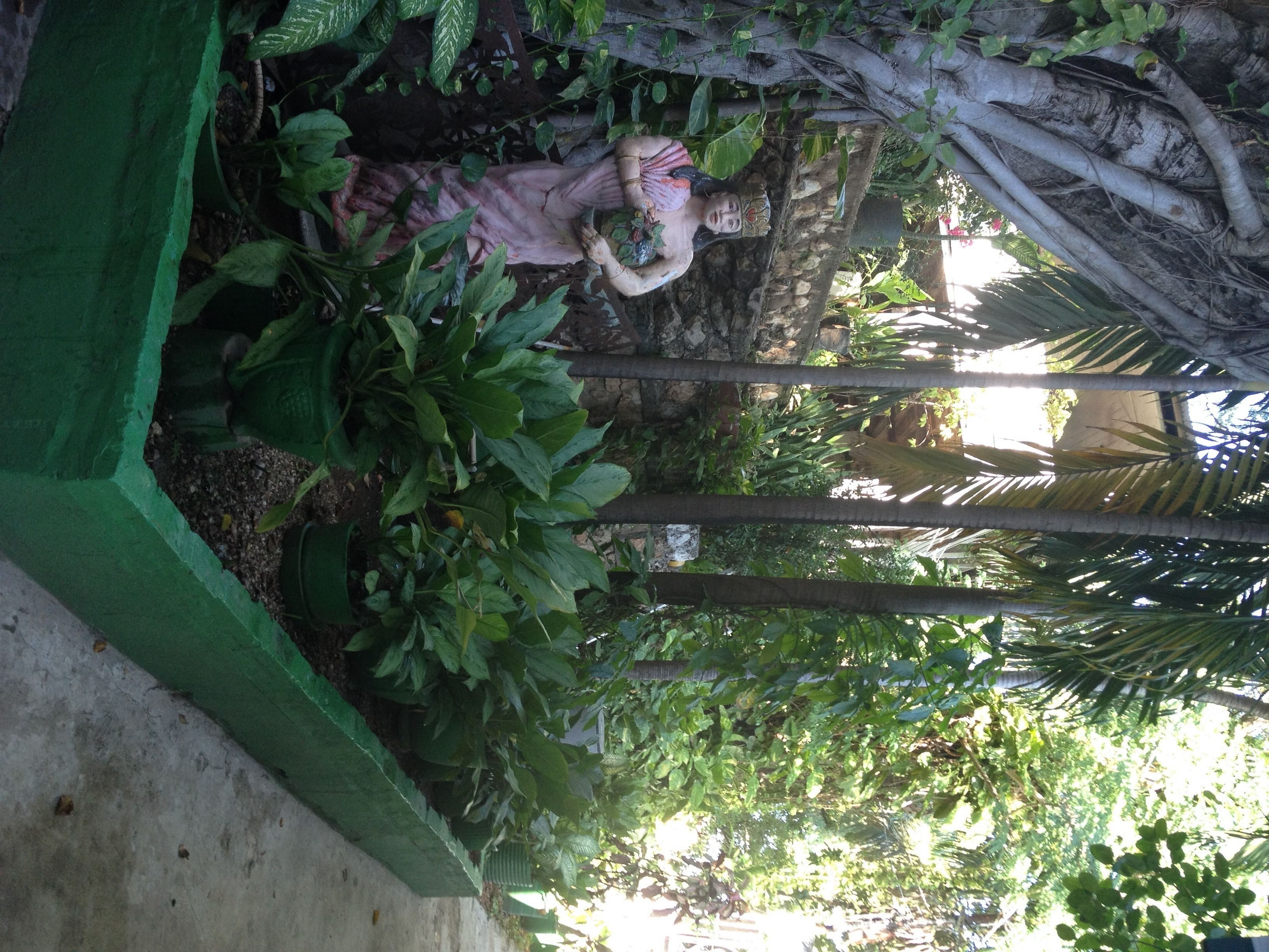 A statue of a woman in a garden with lush greenery and a stone wall.