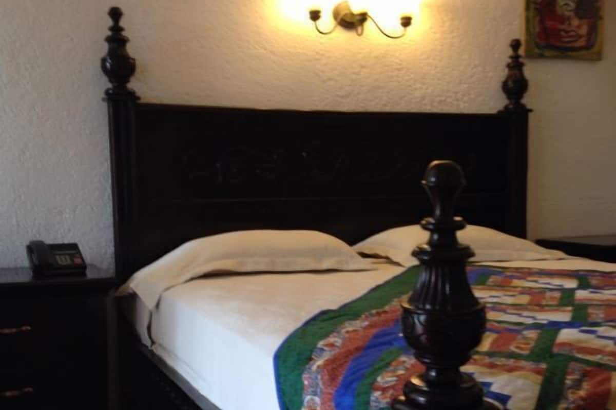A bedroom with a wooden bed, white linens, a quilt, a nightstand, and a wall-mounted light fixture.