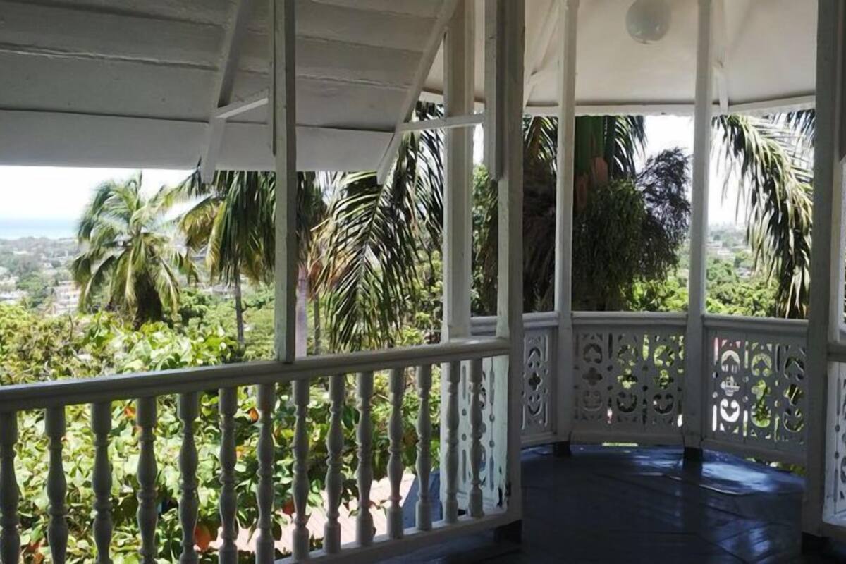 A covered outdoor area with a view of a lush landscape, including palm trees and a distant view of a town.