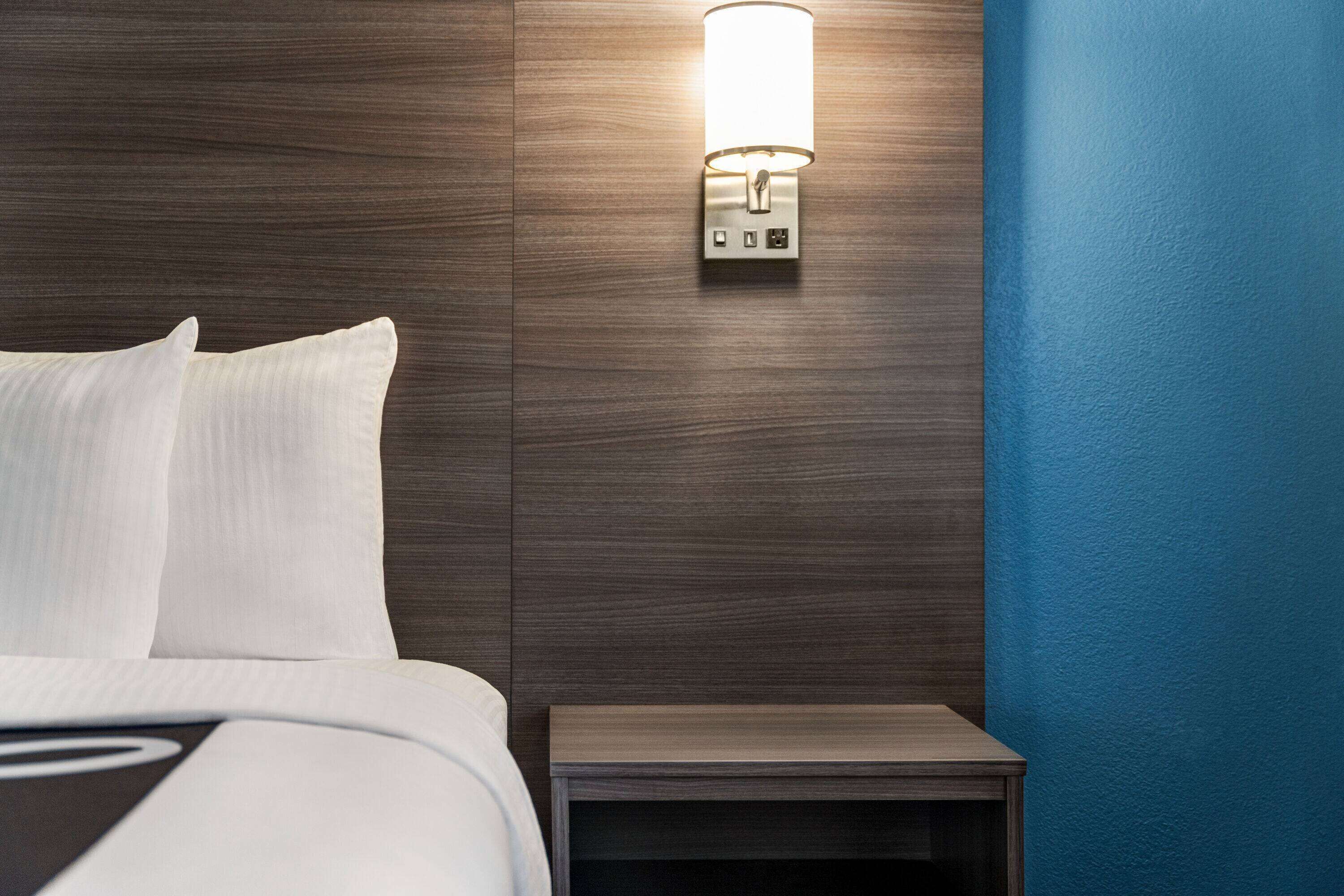 A neatly made bed with white linens, a wall-mounted light fixture, and a small wooden nightstand.