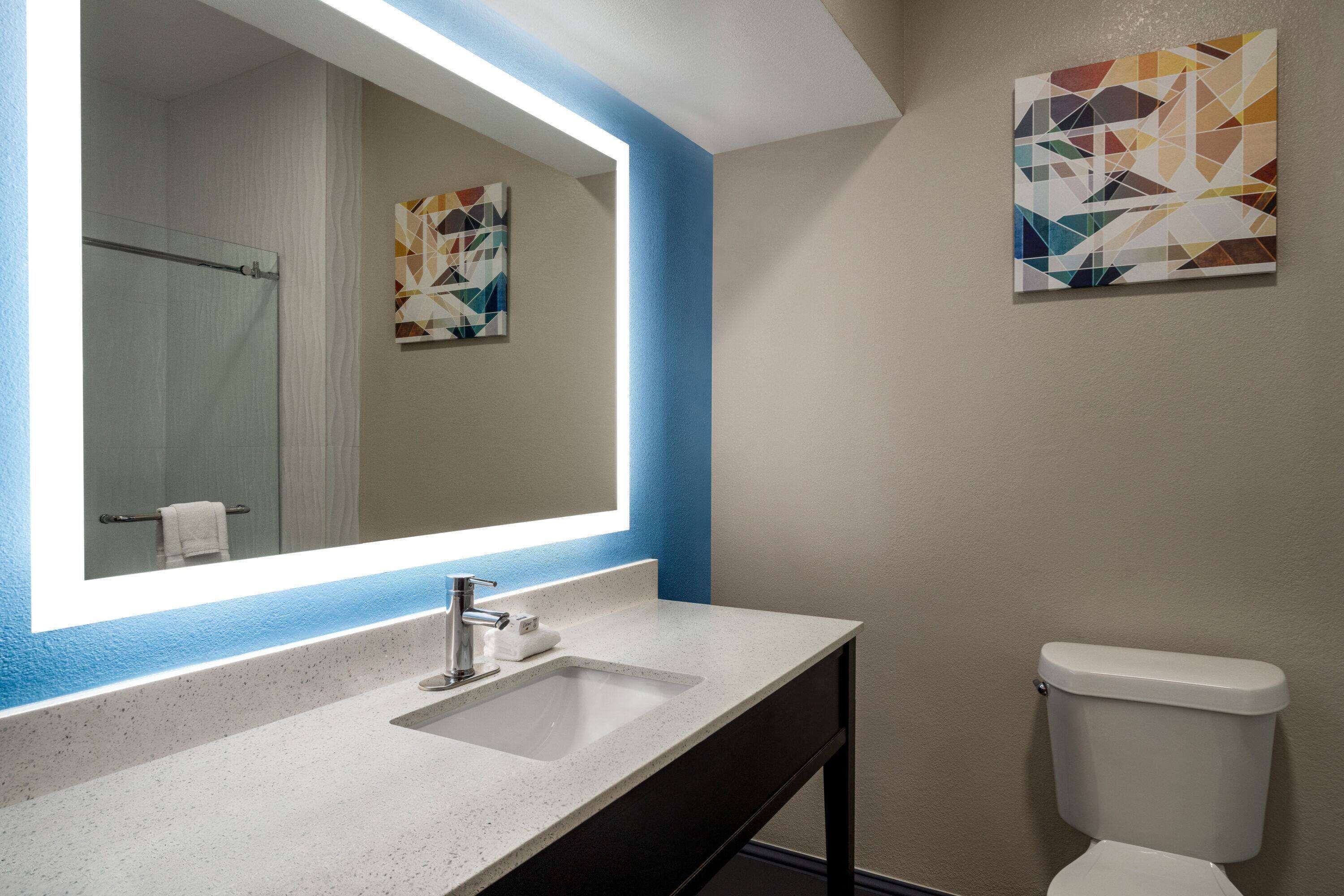 Une salle de bain avec un lavabo blanc, un miroir, des toilettes et deux tableaux abstraits accrochés aux murs.