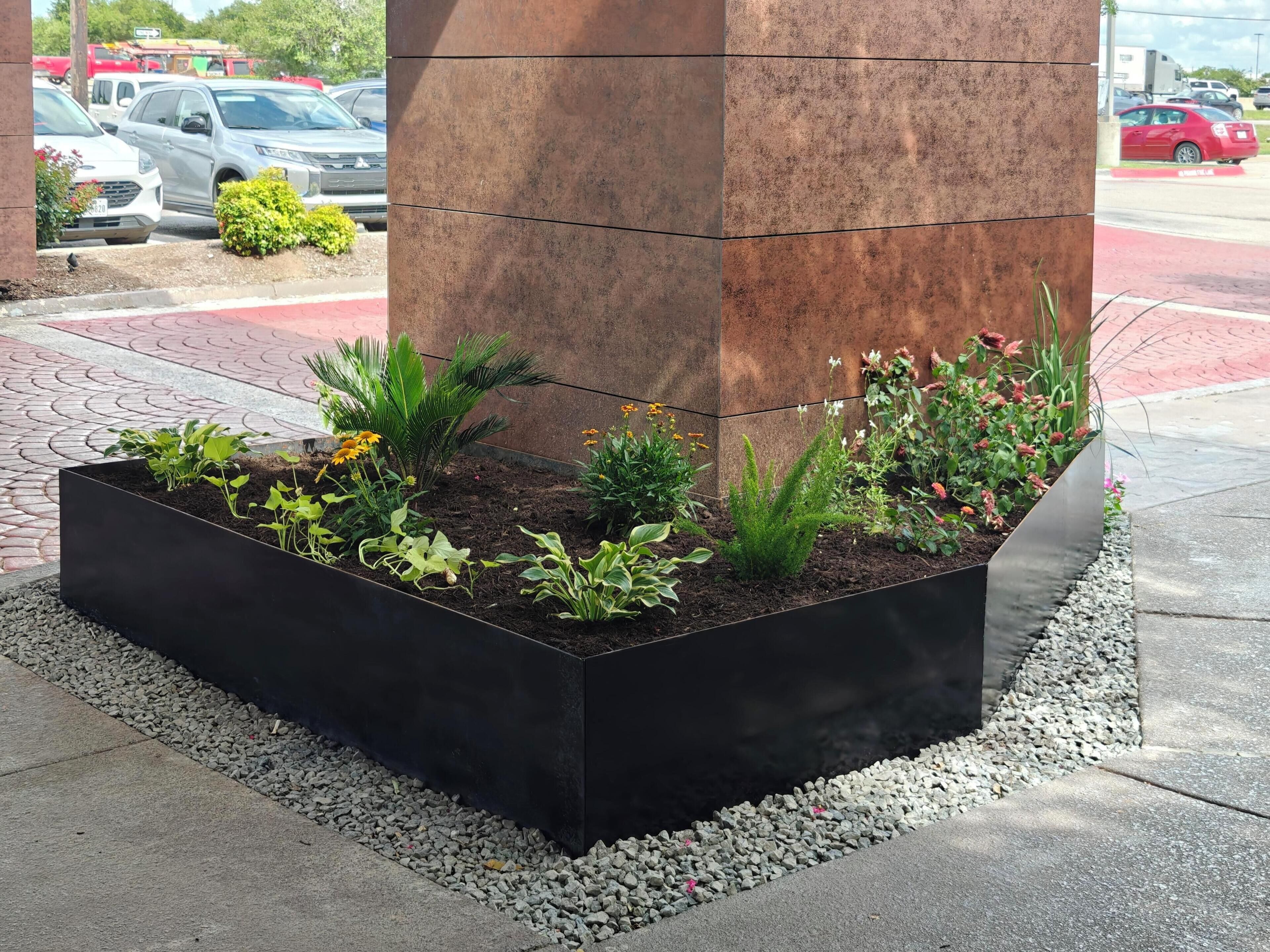 A landscaped planter box with various plants and flowers, positioned against a building's corner.