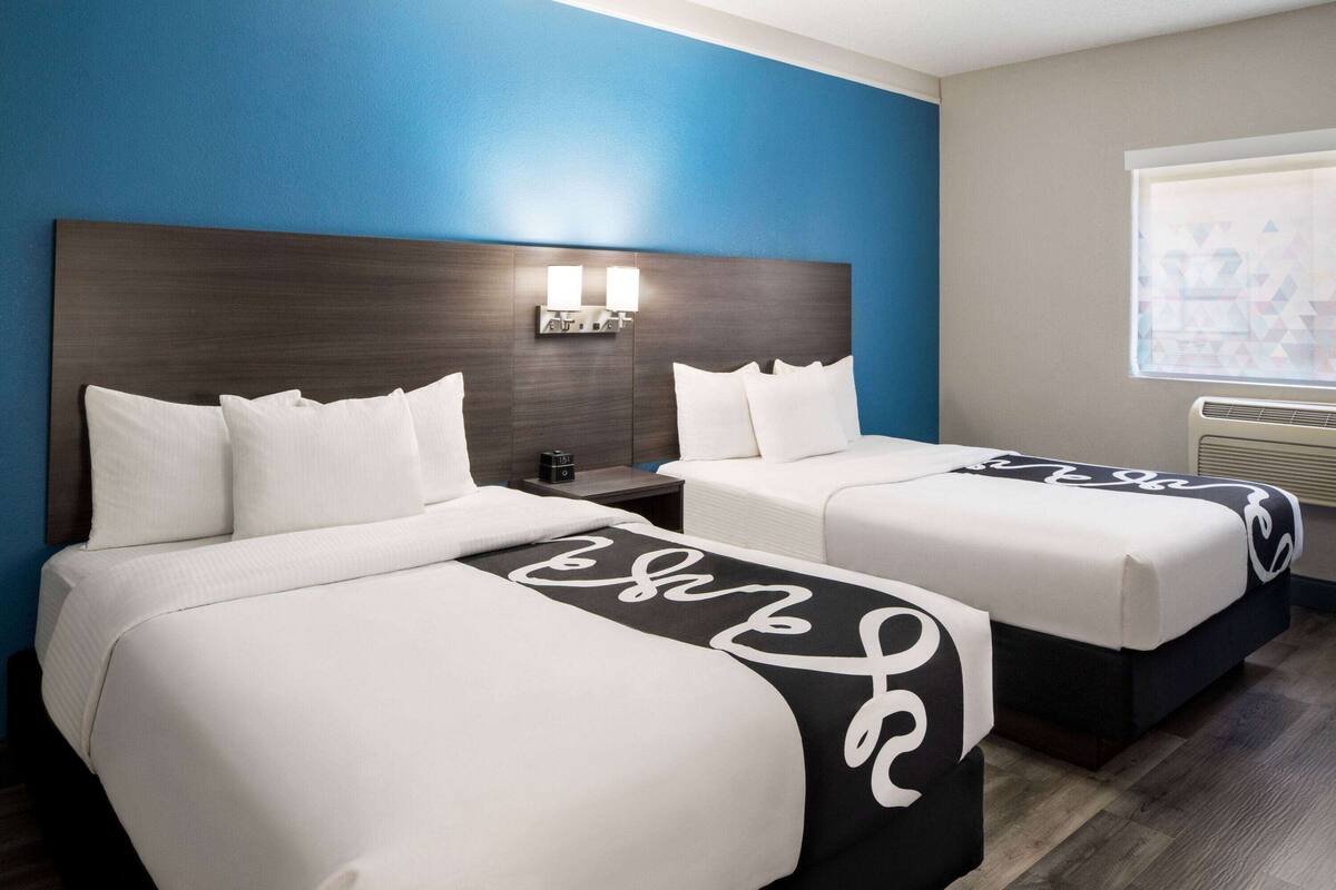 Une chambre d’hôtel avec deux lits, une tête de lit sombre, une table de chevet avec une lampe et une fenêtre avec des stores.