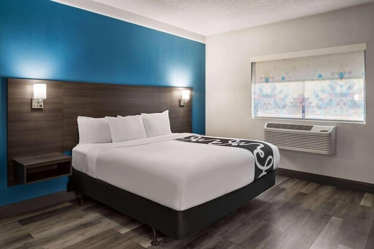 Une chambre d’hôtel moderne avec un grand lit, deux lampes de chevet, une fenêtre avec des stores à motifs géométriques et un climatiseur fixé au mur.