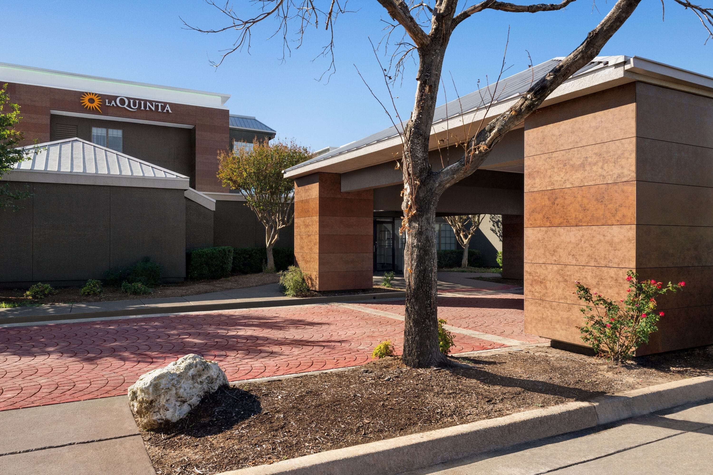 A La Quinta hotel entrance with a brick pathway, a tree, and a rock.