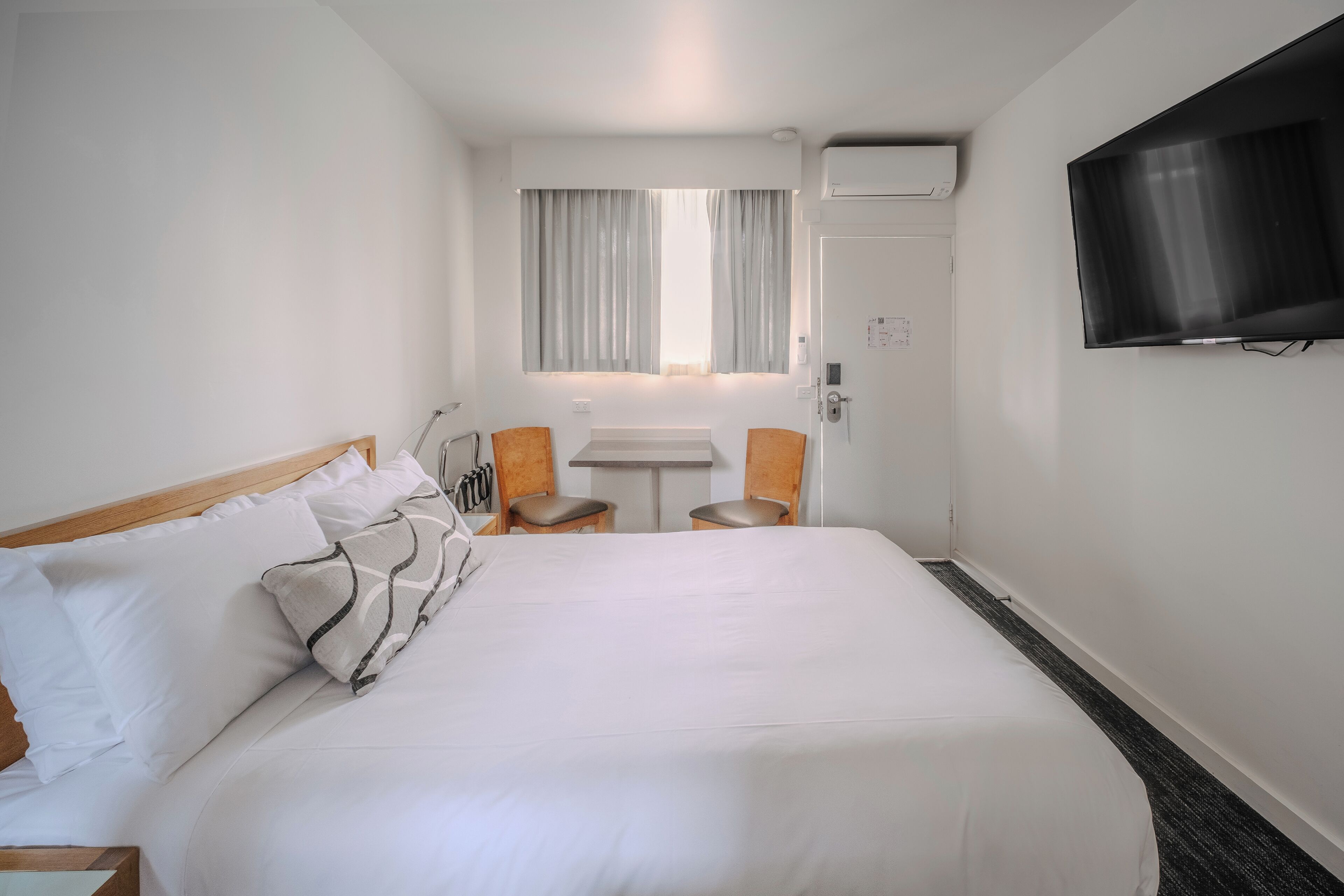 Habitación de hotel con una cama grande, una mesita, dos sillas, un televisor y una ventana con cortinas.