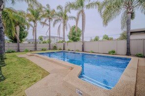 Une piscine rectangulaire dont le bord est recouvert de carreaux, entourée d’une terrasse en béton et d’une zone herbeuse.