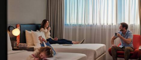 Camera d'albergo con due letti, una zona salotto e un'ampia finestra con tende.