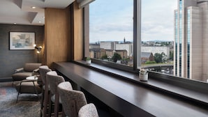 A modern bar area with a long counter, multiple bar stools, and a large window offering a city view.