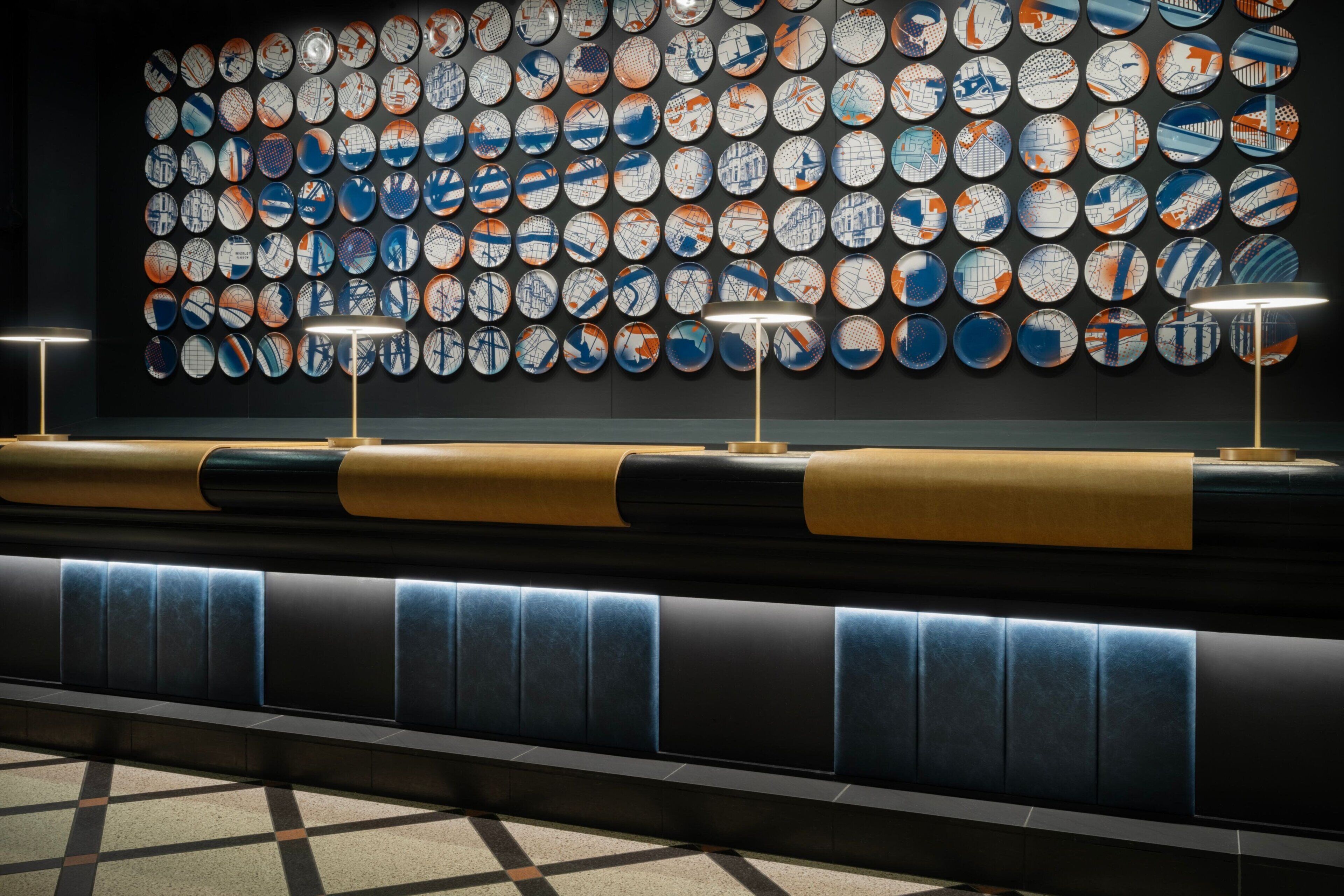 A wall of circular plates with a uniform pattern, a long bench with a patterned backrest, and two floor lamps.