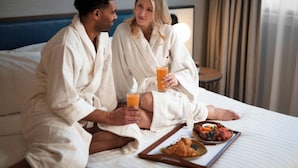 Two people in robes sitting on a bed with a tray of food and drinks.