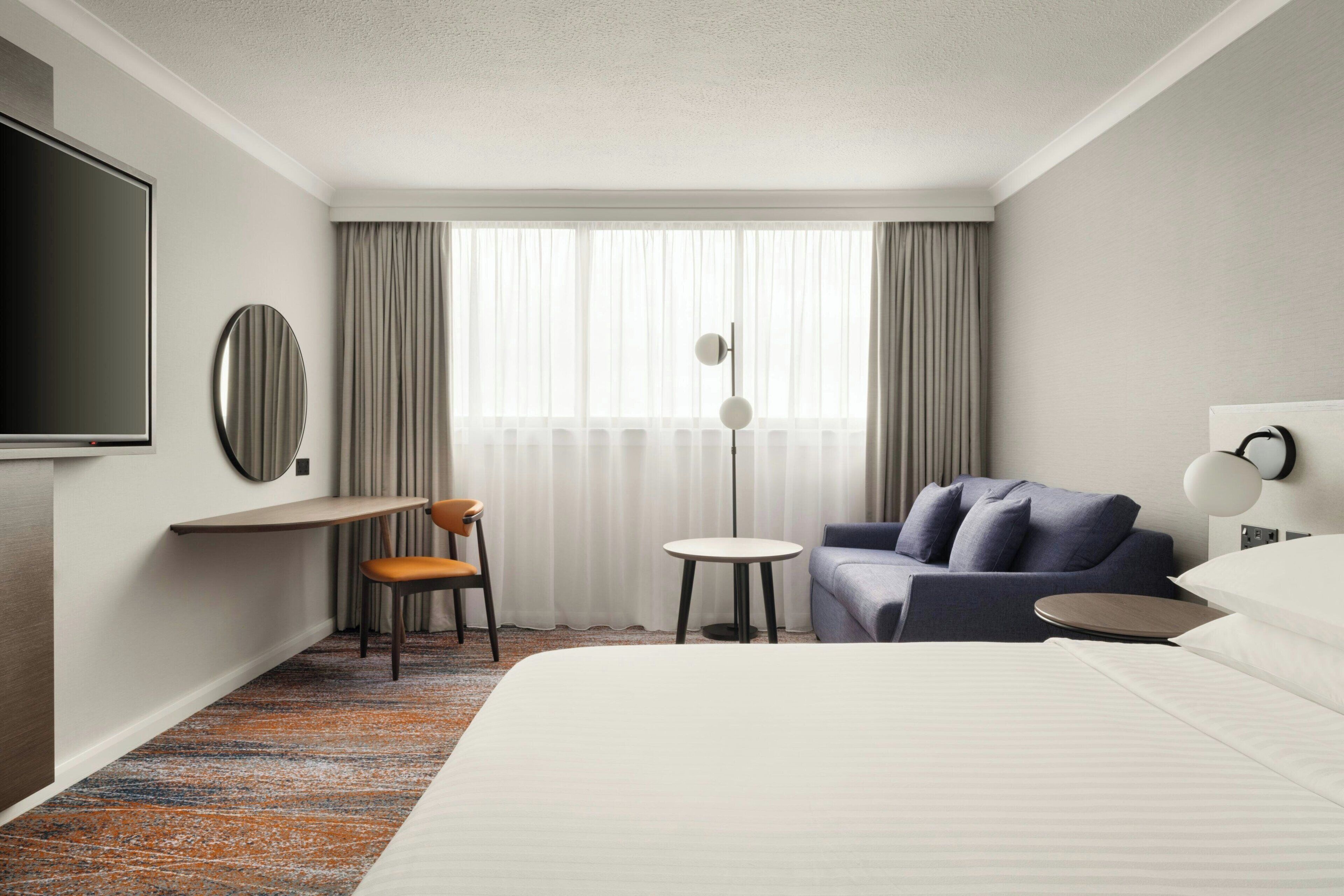 Habitación de hotel con cama, escritorio, silla, sofá, televisión y ventana con cortinas.