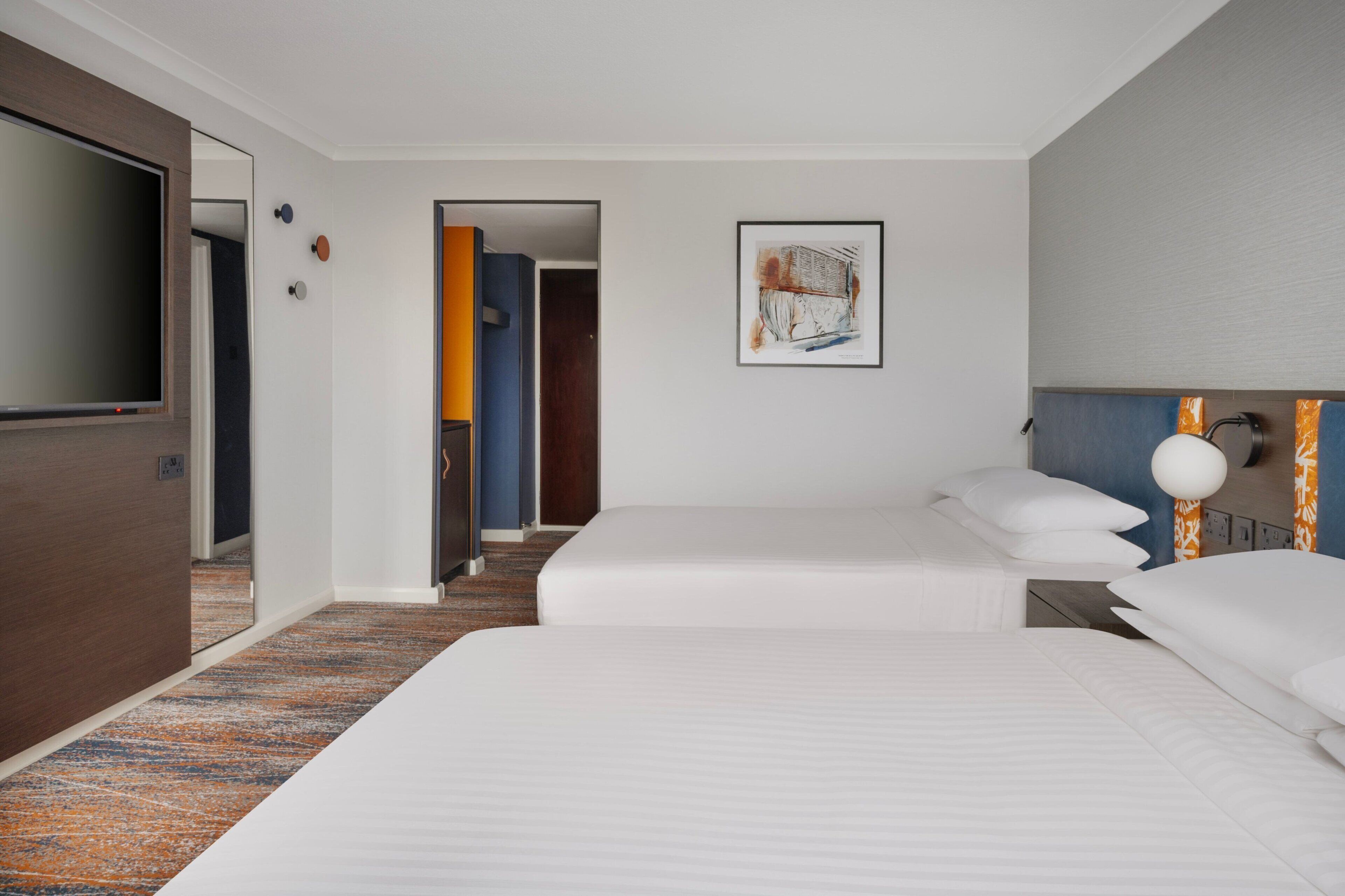 A hotel room with two beds, a television, a framed artwork, and a door leading to another room.