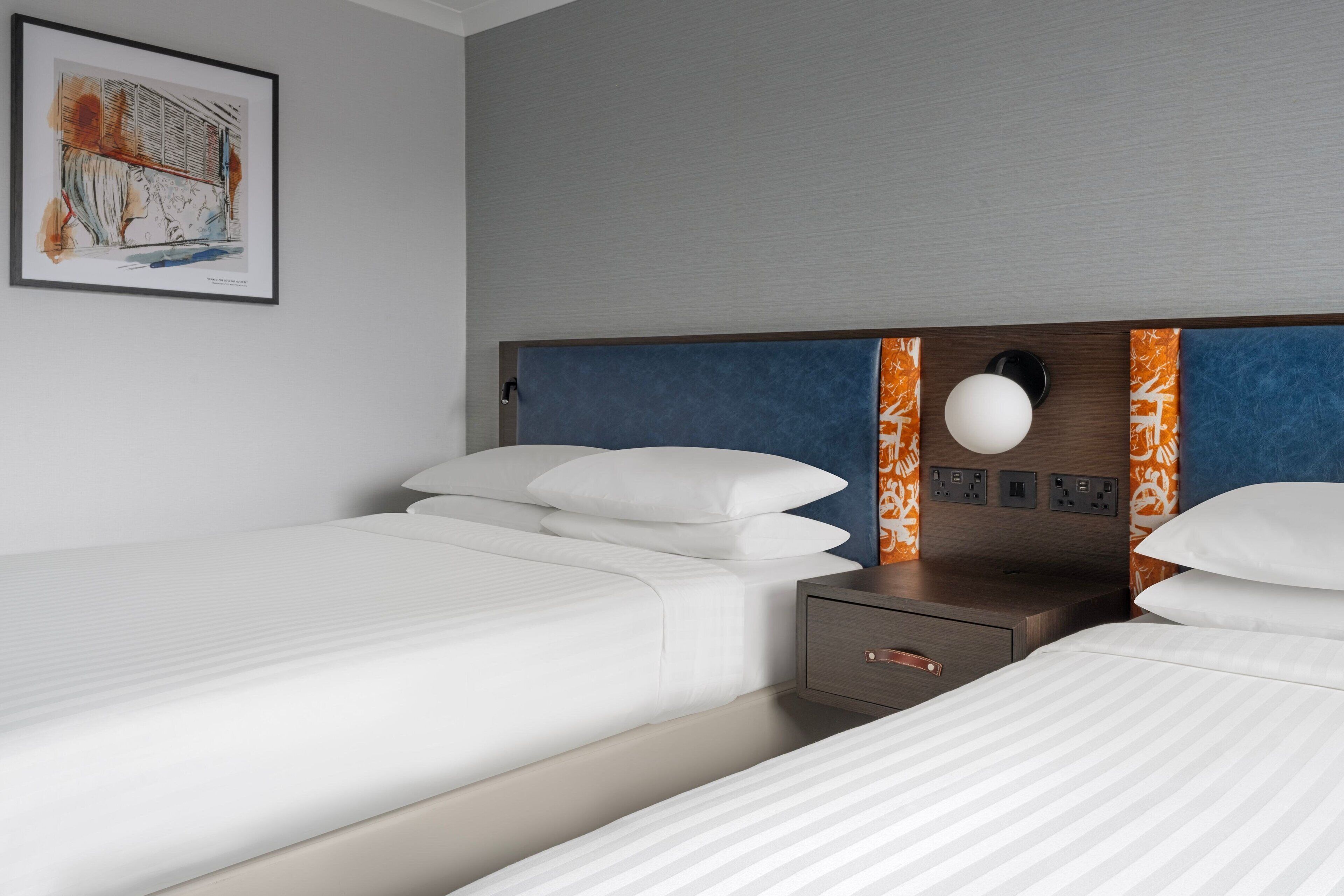 A hotel room with two beds, a nightstand, a wall-mounted lamp, and a framed artwork.