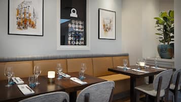 A dining area with two tables, each with four chairs, and a bench. There are framed artworks on the wall, a potted plant, and a window with a view.