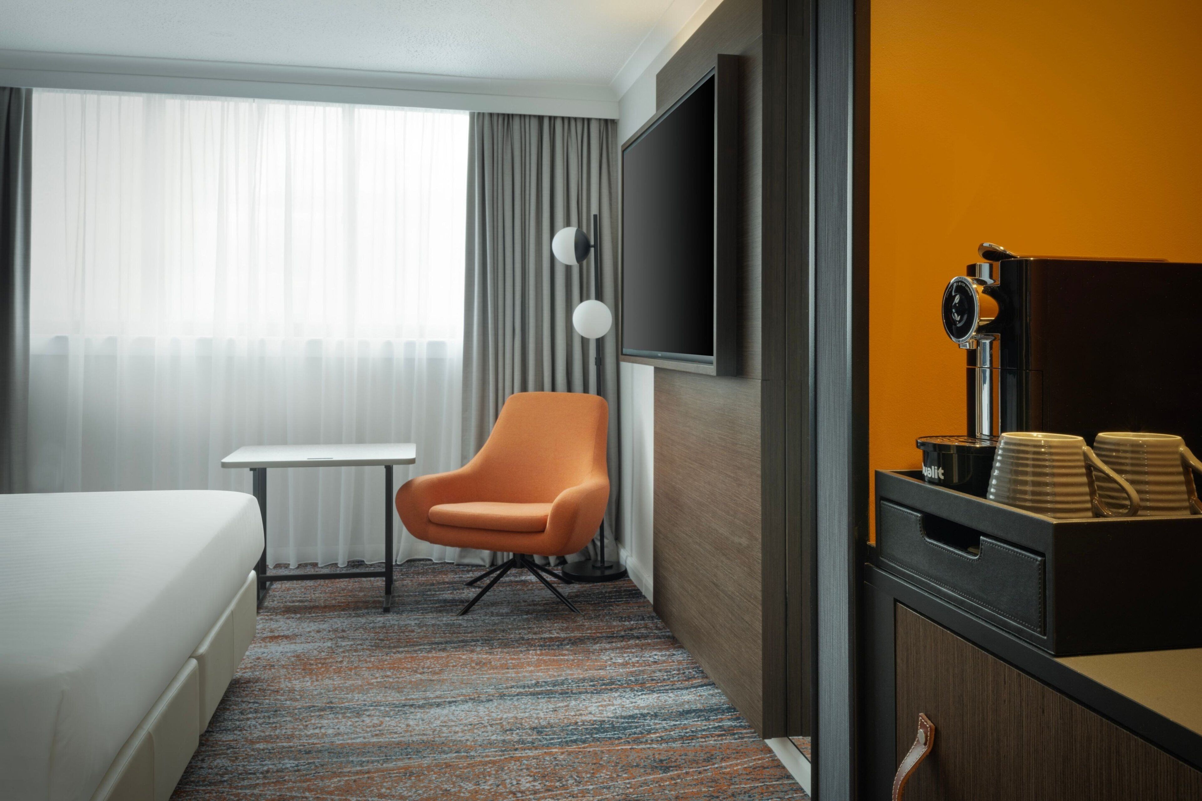 Une chambre d’hôtel équipée d’un lit, d’un bureau, d’un fauteuil orange, d’une télévision et d’un coin café.